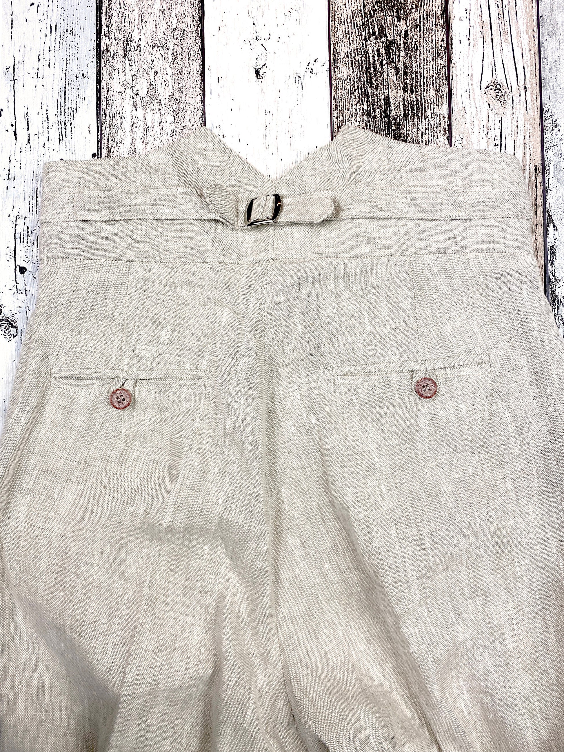 1930s style ladies suit pants for women in natural colored linen. These trousers are adapted from a 1930s mens pattern with wide fit, pleated front, very high waisted, fishtail back and buckle back. Close up view from the back, showing two piped pockets with loop and button closure
