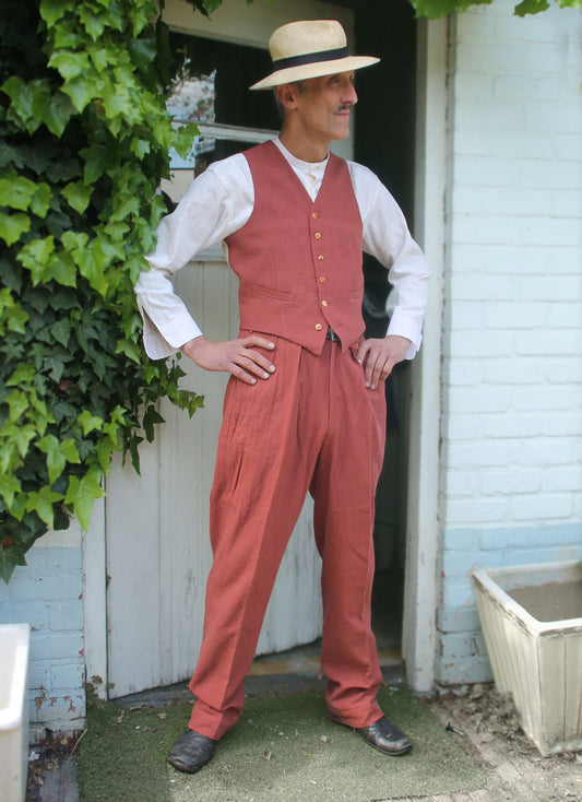 A man wearing a straw hat, white shirt, and a rust-coloured waistcoat and trousers. He is standing in front of a white brick wall with a door behind him.