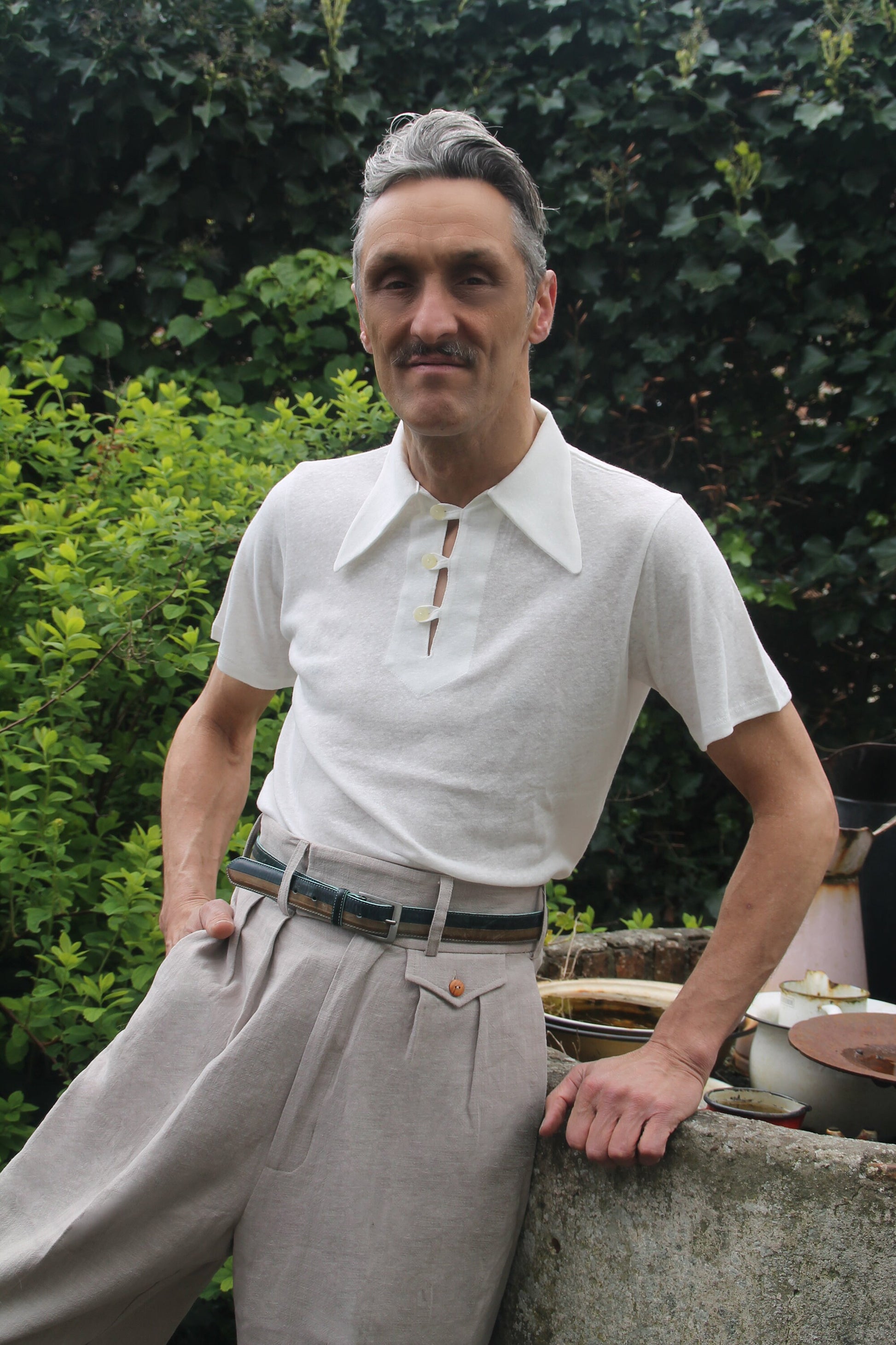 1940s linen high waist pleated trousers with belt hoops and belt, paired with a vintage style off white linen polo shirt. The trousers have belt hoops, inward folded pleats, and a small change pocket with a pocket flap. The colour is light grey