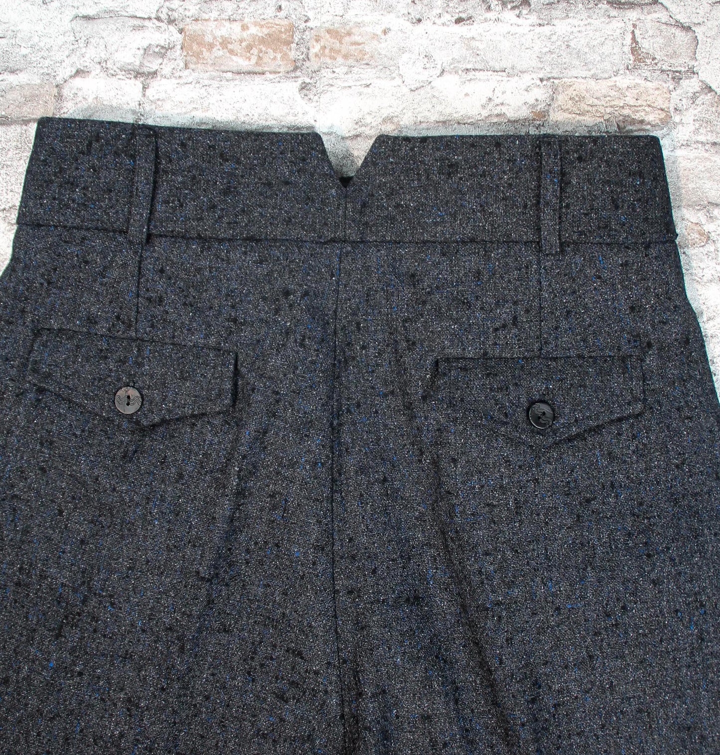 These high-waisted, pleated vintage-style trousers are made of rugged pure wool in a dark grey charcoal colour with small bright blue flecks.  The design is based on an original pattern from the 1940s with all the attention to details. Close up from the back of the trousers, showing two pocket flaps with buttons. The waisband hase a cut out V in the middle,, and two belt hoops
