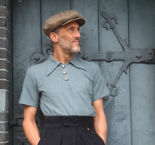 Man wearing a 1930s style sage green polo shirt with buttoned placket, paired with high-waisted dark vintage trousers and a brown flat cap, standing in front of a rustic blue wooden door.