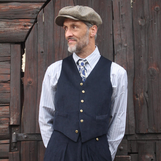 A navy blue waistcoat with gold buttons, worn over a striped shirt and patterned tie. The outfit includes matching navy blue pleated trousers and a tweed newsboy cap. The ensemble is a classic, vintage-inspired look.