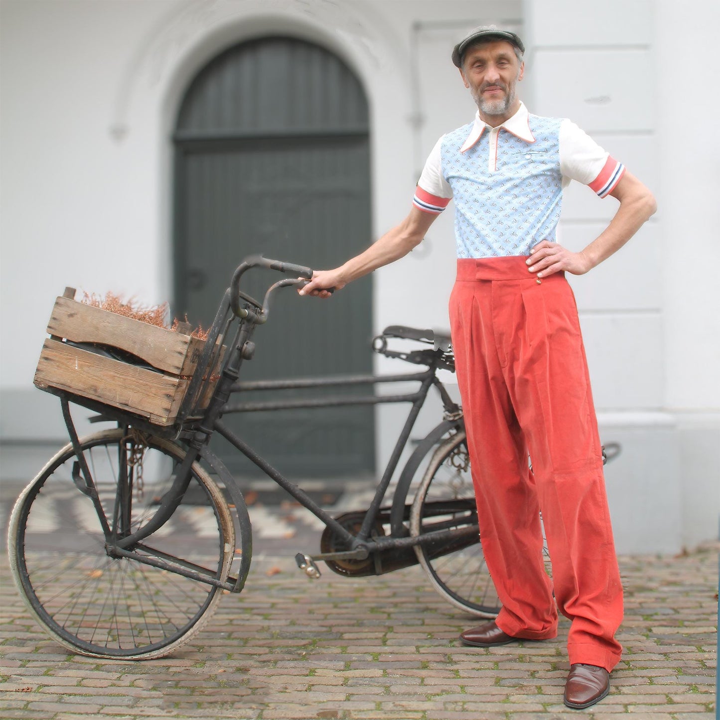 May include: An old black bicycle with a wooden crate. The bicycle is parked on a brick surface. The person is wearing a light blue and white patterned polo shirt, red trousers, and a grey cap.